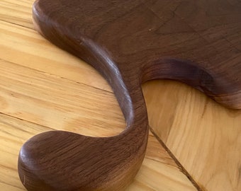 Puede incluir: Una tabla de cortar de madera marrón oscuro con un diseño de mango único. El mango tiene forma de bucle con un extremo redondeado.