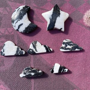 May include: A set of seven black and white marble stones carved into the shapes of a crescent moon, a star, and five clouds. The stones are arranged on a purple fabric background.