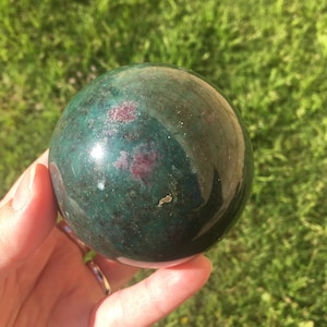 May include: A green and pink gemstone sphere, polished to a smooth finish. The sphere is held in a hand, with the background blurred.