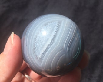 May include: A polished agate sphere with a white and grey banded pattern. The sphere is translucent and has a natural, organic shape.
