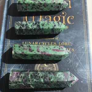May include: Four green, black, and pink gemstone points, also known as zoisite, are arranged vertically on a blue book with gold lettering. The book is titled "A Handbook of Lunar Cycles, Lore, and Magic".