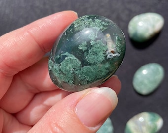 May include: A collection of moss agate tumbled stones. The stones are a mix of green, white, and gray with a translucent appearance.