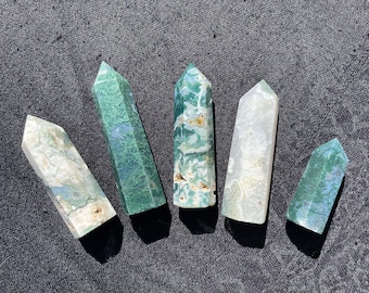 May include: Six green and white agate crystal points, also known as green moss agate, arranged in a semi-circle on a dark grey surface.