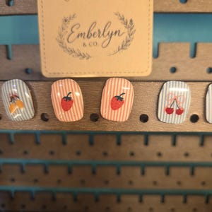 May include: Four rectangular stud earrings displayed on a brown wooden stand. The earrings feature light blue and pink vertical stripes with small fruit illustrations including yellow lemons, red strawberries, and red cherries with a pink bow.