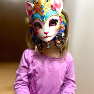 May include: A young person wearing a white, blue, and gold cat mask with intricate details. The mask has a crown and floral accents. The person is wearing a purple long-sleeved shirt with a white unicorn design.