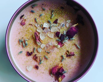 May include: Pink candle with gold glitter, dried rose petals, and white crystals. The candle is in a round, purple-rimmed glass jar.