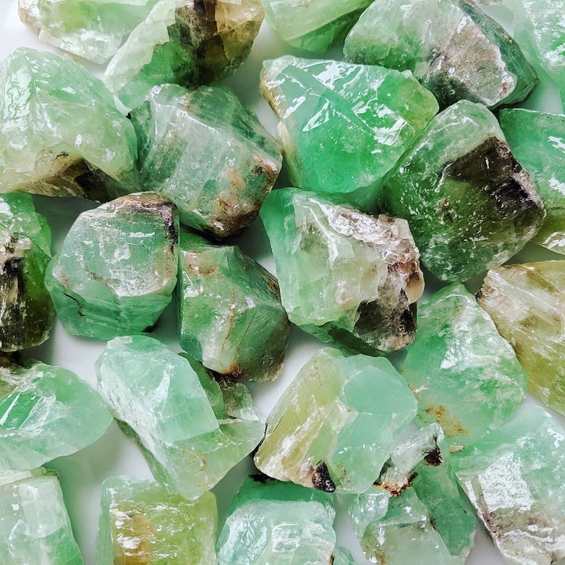 Puede incluir: Primer plano de una colecci&oacute;n de cristales de calcita verde en bruto. Los cristales, de varios tama&ntilde;os y formas, muestran tonos de verde y algunas &aacute;reas marrones y blancas. La textura rugosa y las formaciones naturales son claramente visibles.