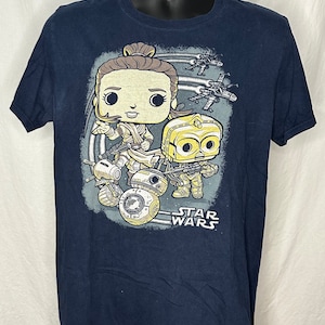 Pode incluir: Uma camiseta azul marinho com um gráfico de Rey e BB-8 de Star Wars em estilo Pop! O gráfico é cercado por um círculo de estrelas e o texto "STAR WARS" é impresso abaixo.