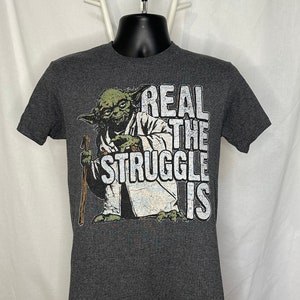 Może przedstawiać: Szary t-shirt z nadrukiem zielonego Yody i tekstem "REAL THE STRUGGLE IS" na przodzie.