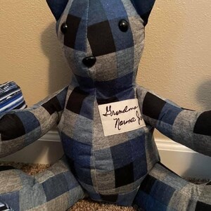 May include: A stuffed bear made from blue, black, and grey plaid fabric. The bear features two round black button eyes and a small black button nose. A white rectangular fabric patch is sewn onto the chest with the text Grandma Norma Jo.
