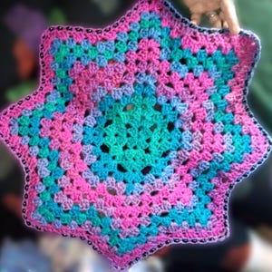 May include: A star-shaped, hand-crocheted item in pink, teal, and purple hues. The edges are trimmed with a darker pink and adorned with small, dark beads. The item is held up by a hand.