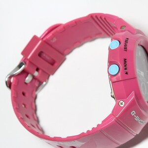 Puede incluir: Un reloj de pulsera digital rosa con una esfera negra y botones azules etiquetados como "MODE" y "ADJUST". El reloj tiene un logotipo G-Shock negro en la parte inferior derecha de la esfera.