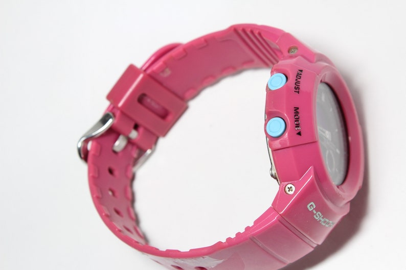 Puede incluir: Un reloj de pulsera digital rosa con una esfera negra y botones azules etiquetados como "MODE" y "ADJUST". El reloj tiene un logotipo G-Shock negro en la parte inferior derecha de la esfera.