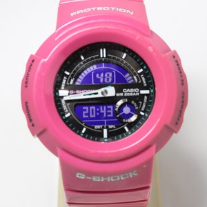 Puede incluir: Un reloj Casio G-Shock rosa con una pantalla digital que muestra la hora 20:43 y una segunda pantalla que muestra el n&uacute;mero 48. El reloj tiene una esfera negra con marcas blancas y una pantalla digital azul. El reloj es resistente al agua hasta 20 bar.