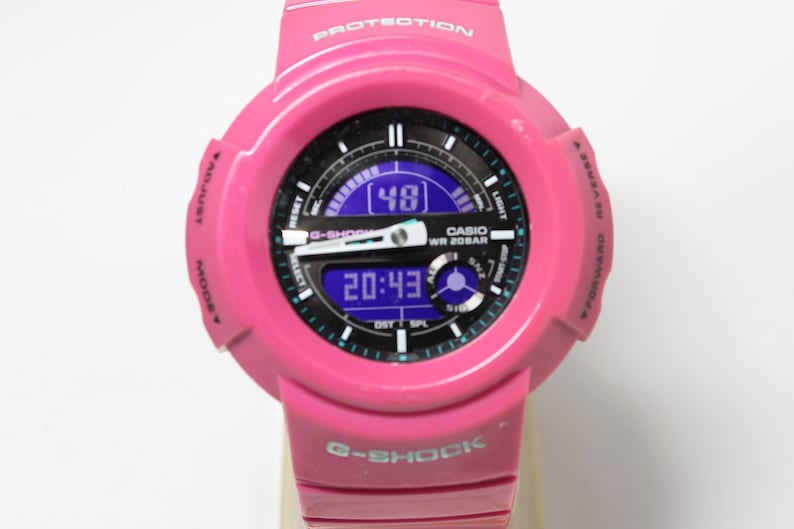 Puede incluir: Un reloj Casio G-Shock rosa con una pantalla digital que muestra la hora 20:43 y una segunda pantalla que muestra el n&uacute;mero 48. El reloj tiene una esfera negra con marcas blancas y una pantalla digital azul. El reloj es resistente al agua hasta 20 bar.