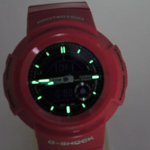 Puede incluir: Un reloj G-Shock rojo con una esfera negra y agujas verdes que brillan en la oscuridad. El reloj tiene una pantalla digital y el texto "PROTECTION" est&aacute; impreso en la parte superior del reloj.
