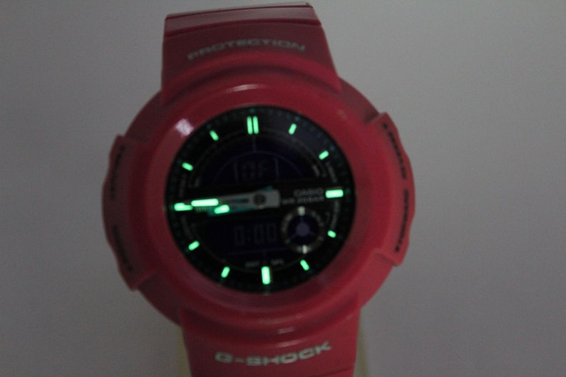 Puede incluir: Un reloj G-Shock rojo con una esfera negra y agujas verdes que brillan en la oscuridad. El reloj tiene una pantalla digital y el texto "PROTECTION" est&aacute; impreso en la parte superior del reloj.