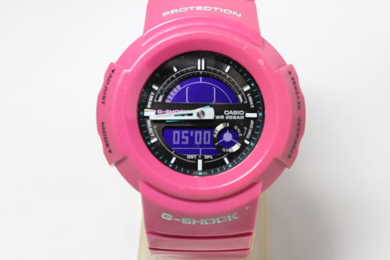 Puede incluir: Un reloj Casio G-Shock rosa con una esfera negra y una pantalla digital. El reloj tiene un acento azul y verde alrededor de la esfera y las palabras "CASIO WR 20BAR" impresas en la esfera.