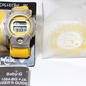 Puede incluir: Un reloj Baby-G plateado con una banda amarilla y una caja negra y amarilla. La esfera del reloj muestra la hora, la fecha y el día de la semana. La caja tiene el texto "Collector by Aunebe" y "Authorized by Information" impreso en ella.