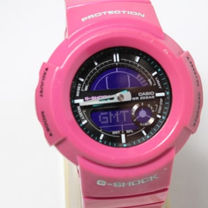 Puede incluir: Un reloj Casio G-Shock rosa con una esfera negra y una pantalla digital. El reloj tiene una correa negra y un logotipo G-Shock blanco en la parte inferior.