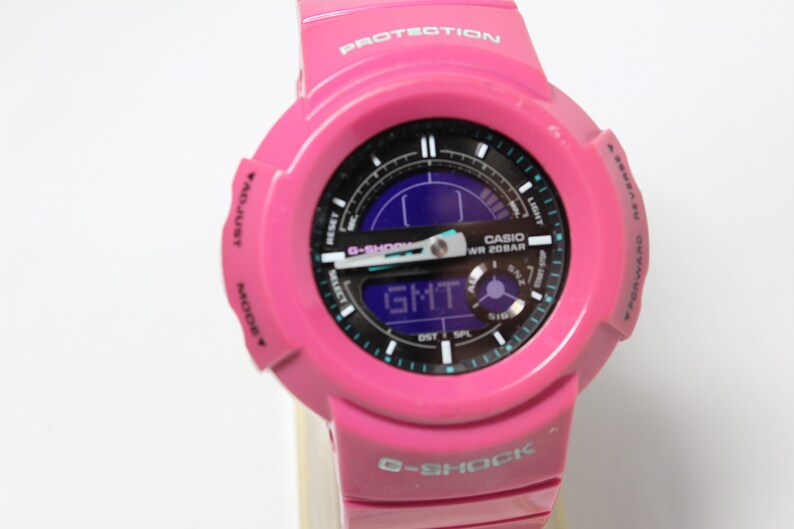 Puede incluir: Un reloj Casio G-Shock rosa con una esfera negra y una pantalla digital. El reloj tiene una correa negra y un logotipo G-Shock blanco en la parte inferior.