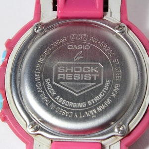 Puede incluir: Un reloj Casio G-Shock rosa y blanco con una parte posterior plateada. La parte posterior del reloj tiene el texto "WATER RESIST 20BAR (4737 AW-582SC ST.STEEL BACK JAPAN MOV'TY CASED IN THAILAND SHOCK ABSORBING STRUCTURE SHOCK RESIST G CASIO"
