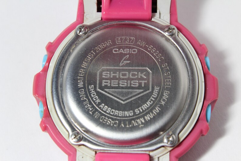 Puede incluir: Un reloj Casio G-Shock rosa y blanco con una parte posterior plateada. La parte posterior del reloj tiene el texto "WATER RESIST 20BAR (4737 AW-582SC ST.STEEL BACK JAPAN MOV'TY CASED IN THAILAND SHOCK ABSORBING STRUCTURE SHOCK RESIST G CASIO"