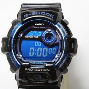 May include: Black and blue G-Shock digital watch with a blue digital display showing the time. The watch face includes the text "G-SHOCK", "CASIO", "WR20BAR", "WORLD TIME", "ILLUMINATOR", and "PROTECTION".