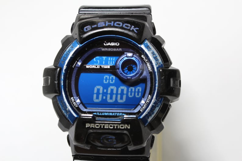 May include: Black and blue G-Shock digital watch with a blue digital display showing the time. The watch face includes the text "G-SHOCK", "CASIO", "WR20BAR", "WORLD TIME", "ILLUMINATOR", and "PROTECTION".