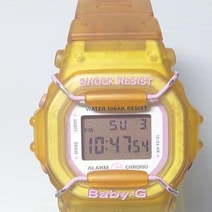 Puede incluir: Un reloj digital Baby-G amarillo con una correa transparente. La esfera del reloj muestra la hora, la fecha y otras funciones. El texto "SHOCK RESIST" y "WATER 10BAR RESIST" es visible. El reloj tiene detalles en rosa.