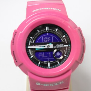 Puede incluir: Un reloj Casio G-Shock rosa con una esfera negra y una pantalla digital. El reloj tiene un acento azul y blanco alrededor de la esfera y las palabras "WR 20BAR" y "PROTECTION" est&aacute;n impresas en el reloj.