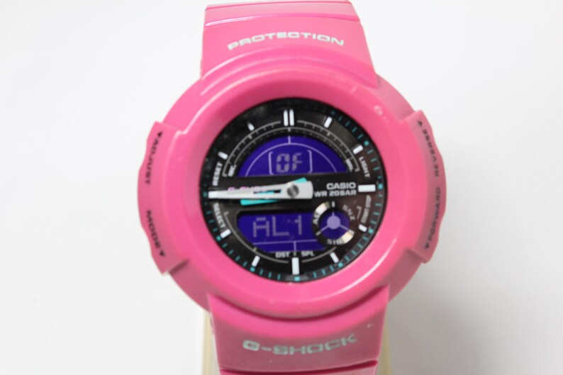Puede incluir: Un reloj Casio G-Shock rosa con una esfera negra y una pantalla digital. El reloj tiene un acento azul y blanco alrededor de la esfera y las palabras "WR 20BAR" y "PROTECTION" est&aacute;n impresas en el reloj.