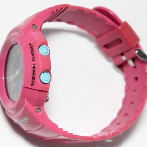 Puede incluir: Un reloj digital rosa con una esfera azul y dos botones azules que dicen "Forward" y "Reverse".