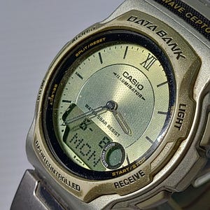 May include: A silver-tone Casio Data Bank Wave Ceptor watch with a gold-colored analog dial and a digital display window. The watch face features the text Casio, Illuminator, Water 5Bar Resist, and Data Bank. It includes buttons labeled Adjust, Split/Reset, Select, Light, Start/Stop, and Receive.