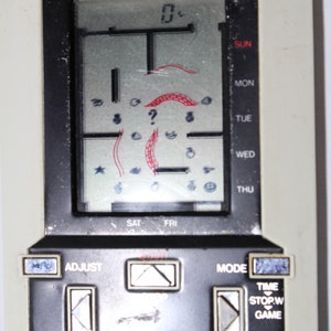Puede incluir: Una consola de juegos portátil en blanco y negro con una pantalla gris que muestra un juego llamado "Pok-Pok man". La pantalla muestra un laberinto con un personaje en el centro. La consola tiene botones para ajustar, fecha, reinicio de vuelta, modo, hora, parada/w, juego y alarma.