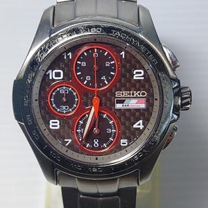 May include: A Seiko BAR Honda chronograph watch with a dark carbon fiber dial, three red-rimmed sub-dials, and a metallic gray stainless steel link bracelet. The watch face features white Arabic numerals, a tachymeter bezel, and a date display window.
