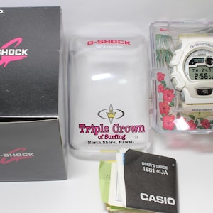 May include: A white G-Shock digital watch with a black and white display, presented in a clear case with red floral accents. The watch is next to a black box with the G-Shock logo and a clear container with the text "Triple Crown of Surfing".