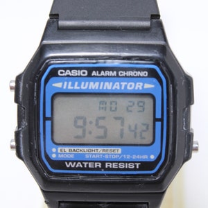 May include: A black Casio Illuminator digital watch with a blue display. The watch face shows the time, date, and day of the week. The watch has a water-resistant feature.