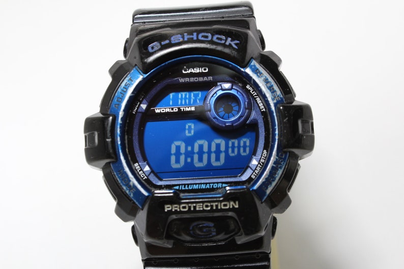 May include: Black and blue G-Shock digital watch with a blue digital display showing 0:00:00. The watch has multiple buttons and features the Casio and G-Shock logos. The watch is designed for protection.