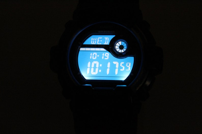 May include: A black digital wristwatch with a bright blue illuminated display. The watch face shows the day, date, and time: Wednesday, 10-19, and 10:17:59. The watch has a round face and a black band.