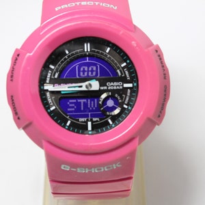 Puede incluir: Un reloj digital Casio G-Shock rosa con una esfera negra y una pantalla digital azul. El reloj tiene una esfera en blanco y negro con el texto "WR 20BAR" y "G-SHOCK".
