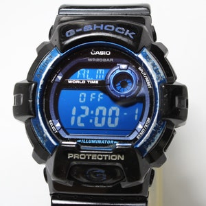 May include: Black and blue G-Shock digital watch with a blue digital display showing the time as 12:00. The watch has a black bezel with blue accents and text that reads "G-SHOCK", "CASIO", and "PROTECTION".