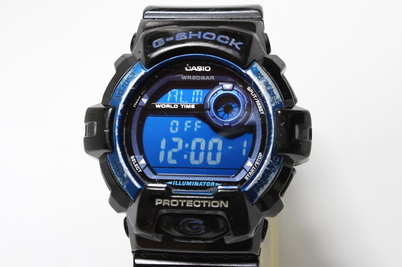 May include: Black and blue G-Shock digital watch with a blue digital display showing the time as 12:00. The watch has a black bezel with blue accents and text that reads "G-SHOCK", "CASIO", and "PROTECTION".