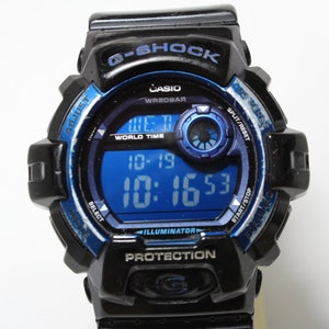 May include: Black and blue Casio G-Shock digital watch. The watch face displays the time and date in a blue digital display. The watch has buttons for adjusting settings and a black band.