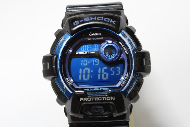 May include: Black and blue Casio G-Shock digital watch. The watch face displays the time and date in a blue digital display. The watch has buttons for adjusting settings and a black band.