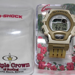 May include: A gold and white G-Shock watch with a digital display and a black strap. The watch is in a clear plastic box with a floral design. The box has the text "Triple Crown of Surfing -26 North Shore, Hawaii" printed on it.