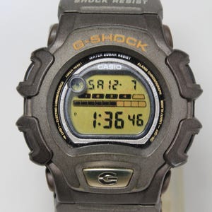 May include: A black and silver G-Shock watch with a camouflage strap. The watch face displays the time, date, and other functions. The watch has the text "SHOCK RESIST", "G-SHOCK", "WATER SUBAR RESIST", "CASIO", "ALARM CHRONOGRAPH", "ON-OFF-LIGHT/ALARM", "ETHNOG" printed on it.