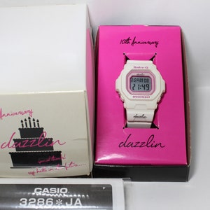 May include: White digital watch with a pink face and pink band. The watch has the text "Baby-G" and "dazzlin" on the face. The watch is in a pink box with the text "10th Anniversary" and "dazzlin".