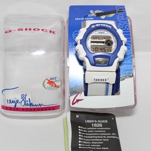 May include: A white and blue G-Shock watch with a digital display and a white and blue striped band. The watch has a black face with a red digital display. The watch is in a clear plastic case with a white and pink label that says "G-Shock" and "Iggie Hakonsen".