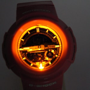 Puede incluir: Un reloj G-Shock rosa con una esfera negra y una luz de fondo naranja brillante. El reloj tiene una pantalla digital y una correa negra.